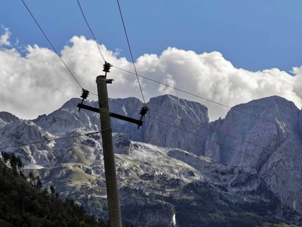 Leaning wooden power poles against rocky mountains and imposing clouds in a bright blue sky, Peaks