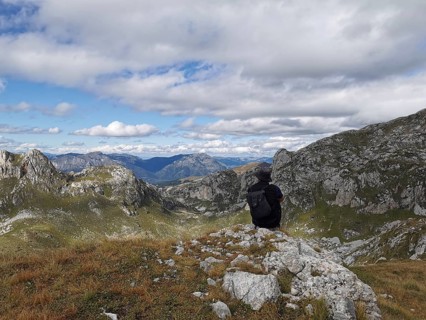 Person with backpack sitting on a rock and looking at a mountain panorama under cloudy sky, Peaks