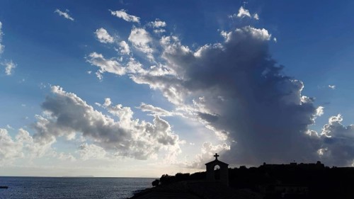 Dramatic sky with clouds and sunlight over sea with church silhouette in foreground, Himare,