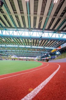 Interior view of a modern sports hall with red running track and green area under steel girders,