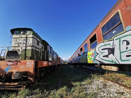 Graffiti-painted locomotive next to abandoned train row, sunny day, Albania
