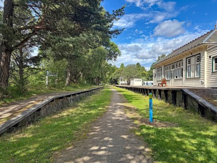 Abandoned station area with wooden buildings and tracks next to a wooded path under blue sky,