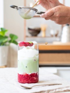 Person sifting green tea powder over a refreshing iced matcha latte with raspberries