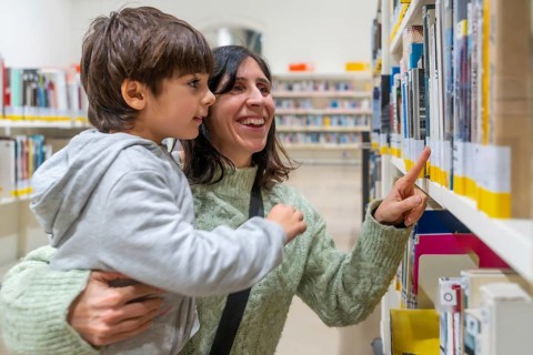 Woman and son smiling together at bookshelves, choosing a book in a library, bonding over reading