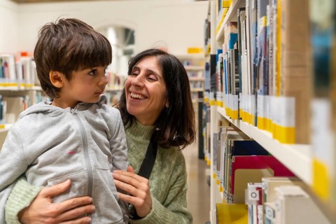 Mother holding her young child, both smiling and interacting happily while standing by shelves
