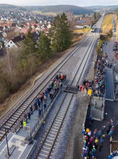 People standing along a railway line next to a small train station, village and countryside