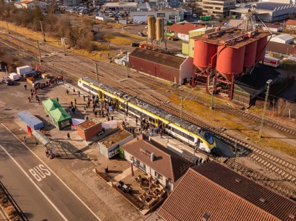Industrial site with a passing train, surrounded by people and various buildings, opening of the