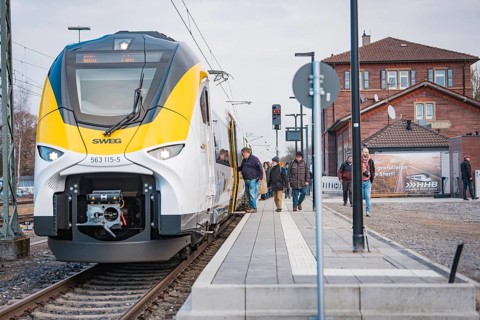 A train on the platform with people and buildings in the background, opening of the Hermann Hesse