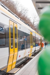 A yellow and white train stands on the platform with people and green balloons, opening of the