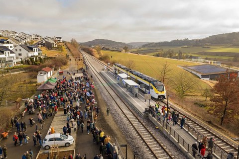 A lively train station in a rural area with lots of people, trains and an idyllic landscape,
