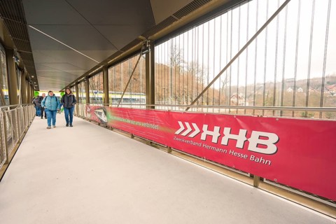 Pedestrian bridge with a large Hermann Hesse Railway banner and people crossing the bridge, opening