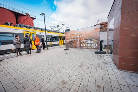 People wait on the railroad track next to a yellow train, a large poster is visible, battery