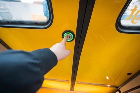 Hand presses button on yellow train door, surrounded by windows, battery electric train Siemens