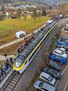 A train arrives in a village, people and parked cars along the tracks, opening of the Hermann Hesse