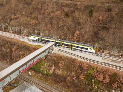 Train on tracks near a bridge with a bare hill in the background and people waiting, opening of the