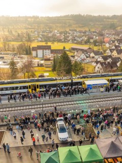 A train with people waiting on a platform in rural surroundings, opening of the Hermann Hesse
