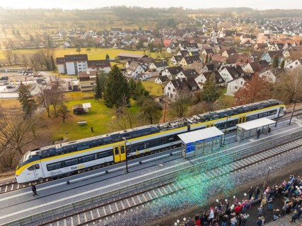 A train is standing at a train station, people are standing to the right, a village and fields in