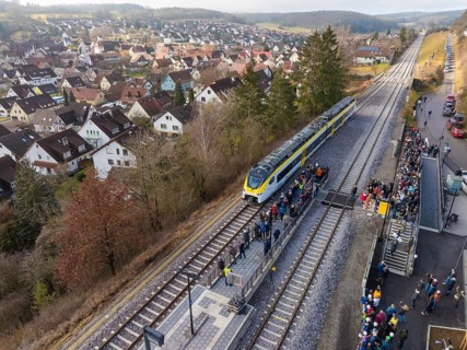 A train stops at a rural train station with surrounding people. The surrounding area shows a