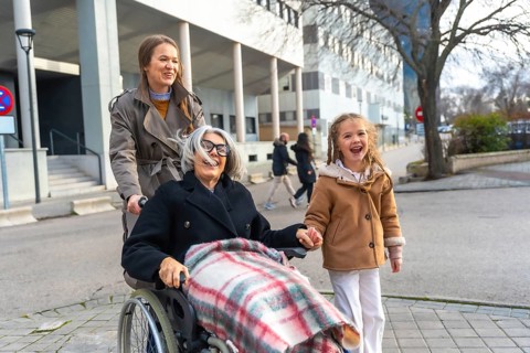 Three generations walking outdoors, smiling grandmother in wheelchair pushed by her daughter while
