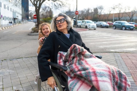 Happy grandchild pushing a senior woman with grey hair and glasses in a wheelchair, enjoying an