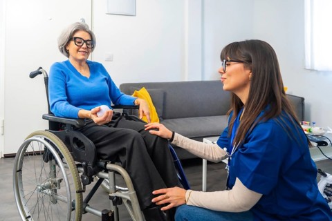 Young female caregiver chatting and smiling with an elderly woman in a wheelchair, offering