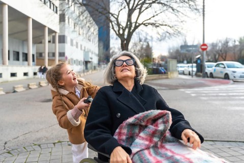 Happy granddaughter pushing senior grandmother in a wheelchair, enjoying a sunny day outdoors and