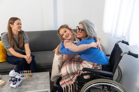 Small girl hugging her grandmother sitting in a wheelchair while a woman watches from the couch,