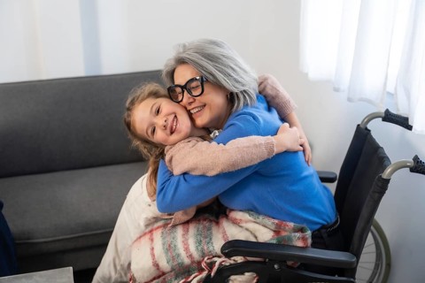 Granddaughter embracing elderly grandmother with gray hair and glasses. Sitting in a wheelchair.