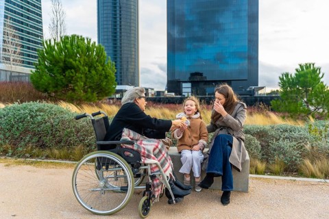 Grandmother in wheelchair enjoys a snack from her smiling granddaughter while family relaxes