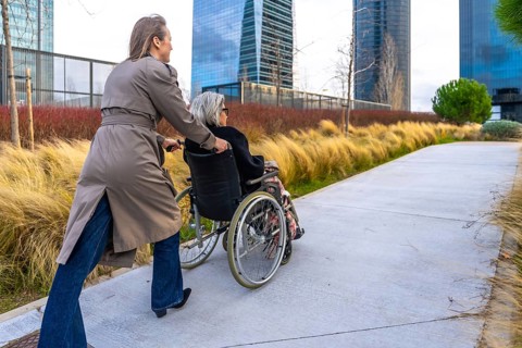 Caregiver pushing an elderly woman in a wheelchair along an outdoor path in a modern city park,