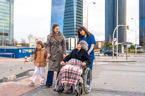 Family members and a professional caregiver are walking outdoors with a smiling senior woman in a