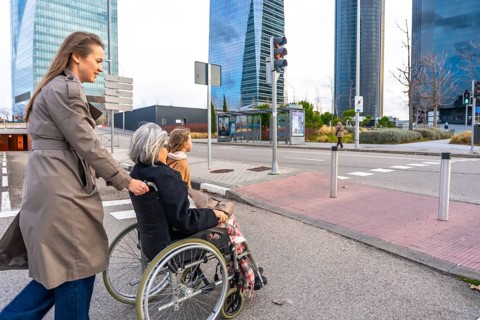 Young woman pushing elderly grandmother in a wheelchair while a child walks beside them across a