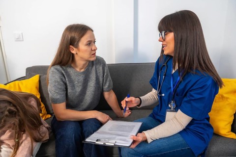 Nurse in scrubs with stethoscope consults a woman at home, explaining medical options and paperwork