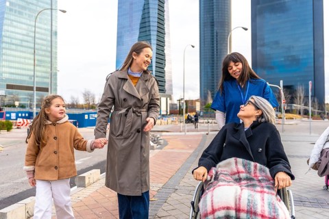Caring caregiver pushing a senior woman in a wheelchair outdoors, joined by her daughter and