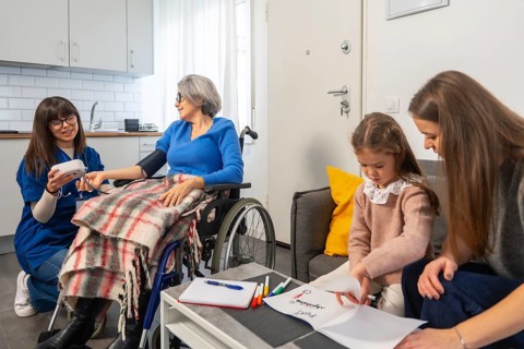 Nurse checks senior woman's blood pressure in wheelchair at home while daughter and grandchild