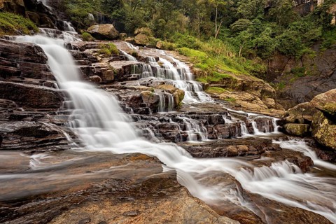 An impressive waterfall flows over stepped rocks surrounded by green nature and thick trees,