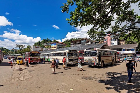 Busy bus stop with people and buses under a sunny sky with green trees, Kandy bus station in Sri