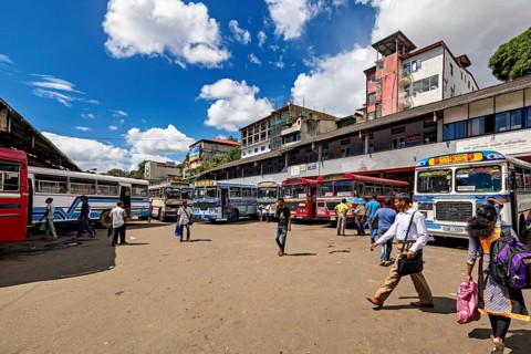 People walk past a bus stop surrounded by various buses and clouds in the sky, Kandy bus station in