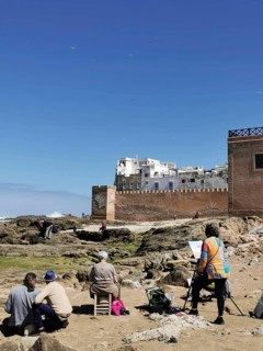 Painters paint the city view on the coast in front of old city walls and rocks under a blue sky,