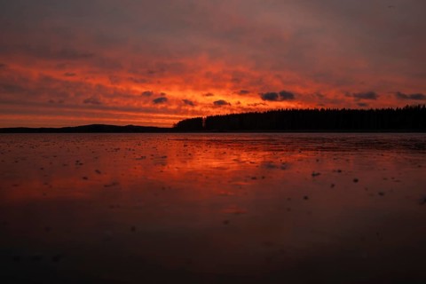 A dramatic sunset over a still, frozen lake and forest with orange-red hues in a cloudy sky,