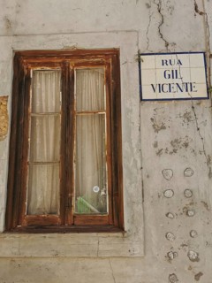 Old window with wooden frame and curtains next to a street sign on a rough, weathered wall