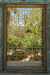 A richly decorated iron window provides a view of a green garden under a clear sky