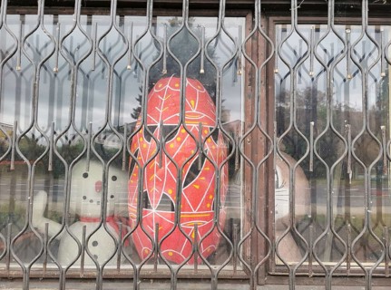 A red abstract sculpture behind a metal grid in a shop window with a street in the background