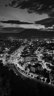 Night view of an illuminated city with mountains in the background under a cloudy sky in black and