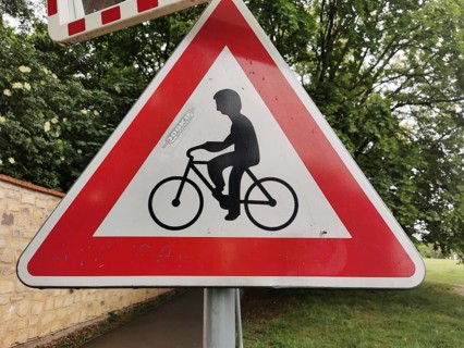 Road sign for bicycles, red and white triangle in the foreground, trees in the background, Czech