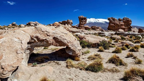 Desert landscape with stone arch and snow-covered mountain in the background under bright blue sky,