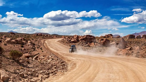 A vehicle drives on a dusty desert road through rocky landscapes under blue skies, The landscape of