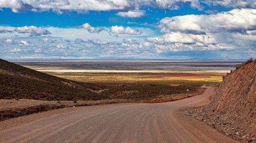 A dirt road leads through a vast, hilly landscape under a cloudy sky, The Landscape of the