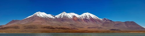Snow-capped mountains above a desert plateau are reflected in a calm lake under a clear blue sky,