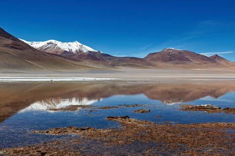 A calm lake reflects snow-capped mountains surrounded by desert-like, open spaces under a clear